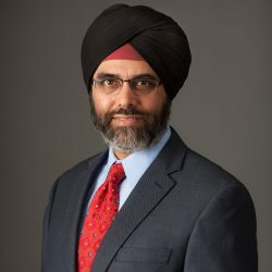 Satwant Dhillon professional headshot wearing a dark suit with a red tie and glasses.