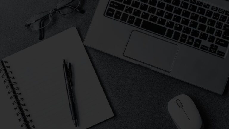 Top-down angled view of a desk with a laptop, notebook, pen, smartphone, coffee, and plant